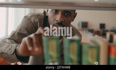 afrokanisch-amerikanischer seriöser, gutaussehender männlicher Student sucht nach Lehrbüchern im Bücherregal in einer großen Bibliothek, die ein Buch nimmt Stockfoto