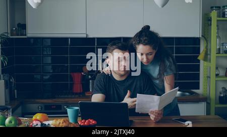Trauriger, unglücklicher junger Mann, der sich Sorgen über die Bezahlung von Rechnungen machte, die mit einem Laptop in der Kithcen zu Hause bezahlt wurden. Seine Frau kommt und unterstützt ihn Stockfoto