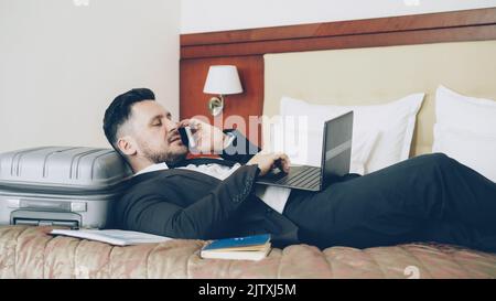 Der junge bärtige Geschäftsmann liegt auf dem Bett und auf dem Koffer, telefoniert und arbeitet von seinem Hotelzimmer aus mit einem Laptop. Reise-, Geschäfts- und Mitarbeiterkonzept Stockfoto