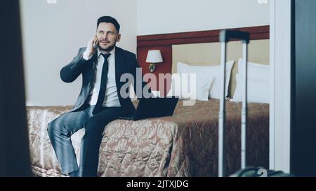 Kippen Sie den lächelnden bärtigen Geschäftsmann in Anzug auf, der am Laptop arbeitet und am Mobiltelefon spricht, während er im Hotelzimmer auf dem Bett sitzt. Business-, Reise- und Mitarbeiterkonzept Stockfoto