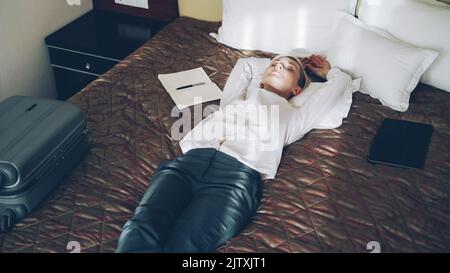 Lächelnde blonde Geschäftsfrau, die entspannt im Hotelzimmer auf dem Bett lag. Reise-, Geschäfts- und Mitarbeiterkonzept Stockfoto