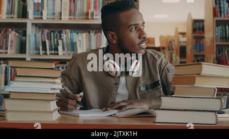 Der positive afroamerikanische junge, gutaussehende Kerl mit großen weißen Kopfhörern sitzt mit Büchern am Tisch, schaut durch das Fenster, lächelt und schreibt in sein Copybook Stockfoto