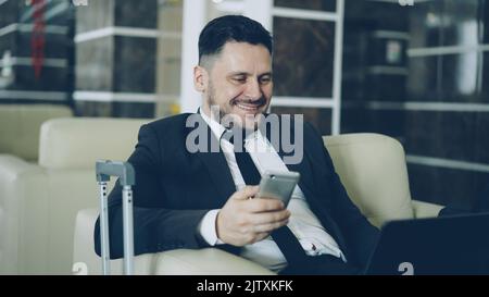 Ein gutaussehender junger Geschäftsmann, der in der komfortablen Hotellobby mit einem Koffer in der Nähe arbeitete und Smartphone und Laptop benutzte. Reise-, Geschäfts- und Mitarbeiterkonzept Stockfoto
