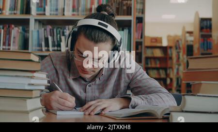 Junger hübscher kaukasischer Student mit großen Kopfhörern sitzt am Tisch in einer großen Bibliothek. Viele Bücher sind um ihn, er lächelt, hört Musik und schreibt im Copybook Stockfoto