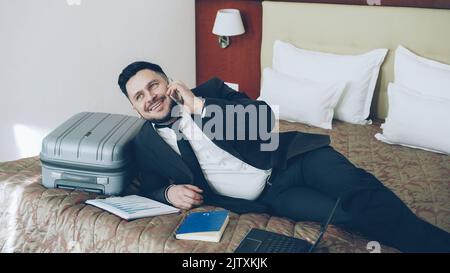 Fröhlicher bärtiger Geschäftsmann, der im Hotelzimmer auf dem Bett lag und über ein Mobiltelefon sprach. Reise-, Geschäfts- und Mitarbeiterkonzept Stockfoto