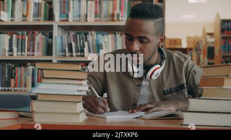 afroamerikanischer junger, gutaussehender Kerl mit großen Kopfhörern sitzt mit Büchern am Tisch und schreibt in seinem Copybook Stockfoto