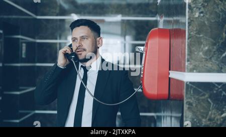 Der bärtige junge Geschäftsmann wählt ein rotes Telefon und spricht mit dem Personal an der Rezeption in der Hotellobby. Business-, Reise- und Mitarbeiterkonzept Stockfoto