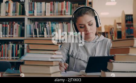 Eine junge lächelnde Frau mit Kopfhörern sitzt am Tisch in der Universitätsbibliothek und hält ein digitales Tablet, hört Musik und schreibt einen Vortrag in einem Notizbuch Stockfoto
