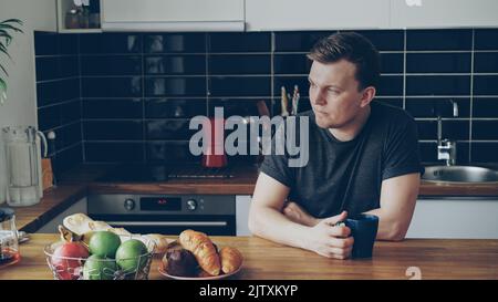 Porträt eines jungen kaukasischen gutaussehenden Mannes, der am Tisch in einer modernen geräumigen Küche sitzt, durch das Fenster schaut, Tee trinkt, ist er müde und aufgeregt Stockfoto