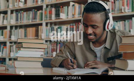 Ein hübscher afroamerikanischer Student, der große weiße Kopfhörer trägt, hört Musik, die am Tisch in einer großen, geräumigen Bibliothek sitzt und einen Vortrag schreibt, der von Büchern umgeben ist Stockfoto