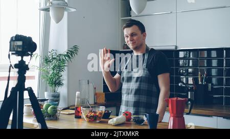 Fröhlicher attraktiver Mann, der Video-Food-Vlog über gesundes Kochen auf einer Digitalkamera in der Küche zu Hause aufzeichnet. Vlogging, Jugendkultur und Social-Media-Konzept Stockfoto