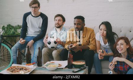 Multiethnische Gruppe von Freunden Sportfans, die sich die Fußballmeisterschaft im Fernsehen anschauen, zusammen Pizza essen und zu Hause Bier trinken Stockfoto