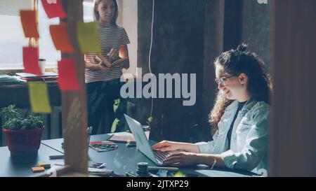 Zwei attraktive Mädchen arbeiten im modernen Loft-Büro im Innenbereich zusammen Curly Businesswoman tippt auf einem Laptop am Tisch, während ihre Kollegin in der Nähe des Fensters ein Buch liest. Stockfoto