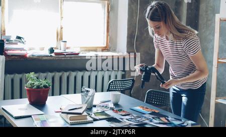 Junge attraktive Bloggerin mit Digitalkamera, um bunte Bilder von Reisen auf dem Tisch angezeigt zu schießen. Sie bewegt Fotos und macht verschiedene Collagen. Büro im Loft-Stil im Hintergrund. Stockfoto