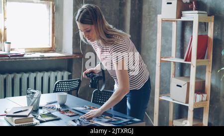 Junge attraktive Bloggerin mit Digitalkamera, um bunte Bilder von Reisen auf dem Tisch angezeigt zu schießen. Sie bewegt Fotos und macht verschiedene Collagen. Büro im Loft-Stil im Hintergrund. Stockfoto