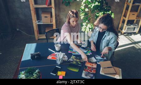 Aus der Perspektive junger Designerinnen werden Fotos auf einen breiten Tisch gelegt, um im Studio eine perfekte flache Lage zu schaffen. Sie saßen zusammen am Schreibtisch und diskutierten über verschiedene Fotos Stockfoto