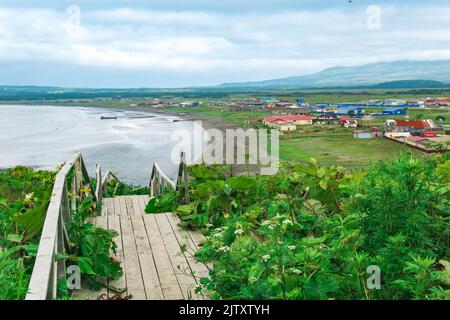 Blick auf die Bucht von Juschno-Kurilsk auf der Insel Kunaschir von einem hohen Kap, im Vordergrund ein hölzerner Bürgersteig mit einer Leiter Stockfoto