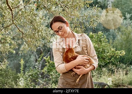 Frau mit brauner Henne in den Händen auf dem Bauernhof. Freiweidende Haushenne auf einem traditionellen Bio-Bauernhof mit freilandem Geflügel. Stockfoto