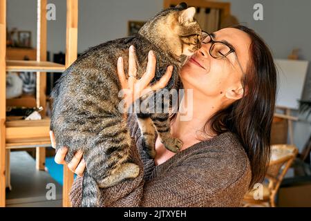 Frau mittleren Alters umarmt niedliche tabby Katze in Indoor-Szene. Mensch-Tier-Beziehungen. Lustige Heimtiere. Heimatlose Haustiere. Stockfoto