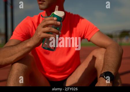 Sportmann macht eine Pause und entspannt sich nach dem Training oder Laufen um das Stadion und Trinkwasser. Sport Lifestyle Konzept. Stockfoto