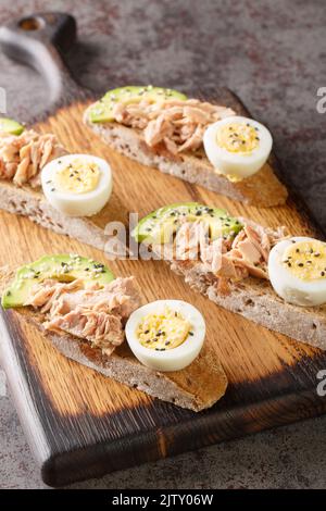 Toast mit Thunfisch in der Dose, gekochtem Ei und reifem Avocado in Nahaufnahme auf einem Holzbrett auf dem Tisch. Vertikal Stockfoto
