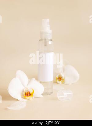 Sprühspenderflasche mit leerem Etikett in der Nähe von weißen Orchideenblüten auf hellbeige Nahaufnahme, kosmetisches Modell. Hautpflege Beauty-Produkt-Paket. Exotisches na Stockfoto