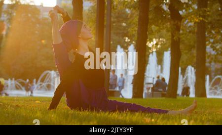 Starke islamische muslimische indische Frau in Hijab aktiv Mädchen tun Yoga im Park im Freien Training Übungen anspannen Muskeln zeigen akrobatische Elemente Stockfoto