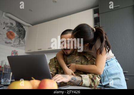 Junge Frau im Küchenzimmer küsst lächelnden Militärmann auf der Wange Stockfoto