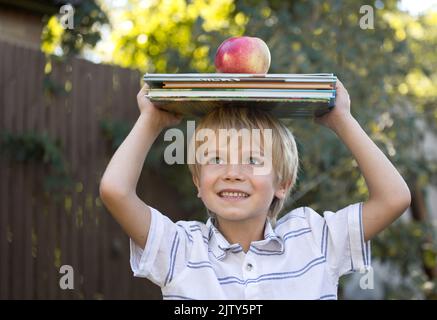 Netter fröhlicher 6-jähriger Junge hält einen Stapel Bücher und einen großen Apfel auf dem Kopf. Bücher lesen, interessante Kindheit, neugieriges Kind, Wissen Stockfoto