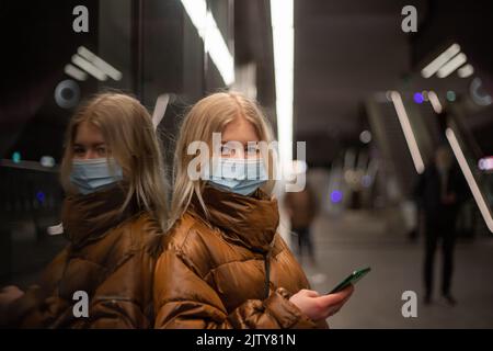 Ein skandinavisches Mädchen trägt eine OP-Maske, während sie auf die U-Bahn wartet und ihr Smartphone nutzt Stockfoto