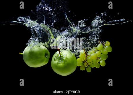 Grüne Äpfel und weiße Trauben fielen in Wasser, wobei Spritzer und Blasen auf schwarzem Hintergrund isoliert wurden. Stockfoto