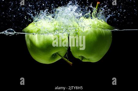 Zwei grüne Äpfel, die mit schwarzen Spritzern in die Wasseroberfläche gelangen. Stockfoto