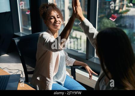 Lächelnde Frauen geben sich im modernen Coworking hohe Fünfen Stockfoto