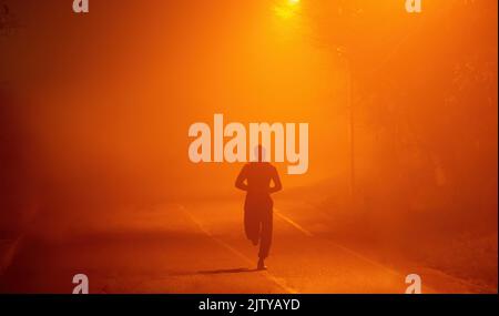 Silhouette eines Läufers im Nebel. Athletischer muskulöser junger Mann, der die mit Rauch gefüllte Straße entlang joggt. Er läuft in der abendlichen städtischen Umgebung in Stockfoto