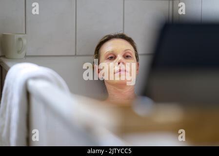 Frau, die sich in einem heißen Bad entspannt, liest auf einem Tablet oder Smartphone mit Fokus auf ihr Gesicht Stockfoto
