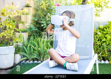 Kleines Mädchen mit Virtual-Reality-Brille, hält sie mit den Händen, blickt nach links und sitzt auf einer Sonnenliege im Garten ihres Hauses. Metav Stockfoto