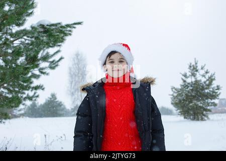 Porträt eines niedlichen Teenagers in einer dunklen Jacke und einem roten Pullover steht im Winter ein roter weihnachtsmann-Hut im Park Stockfoto