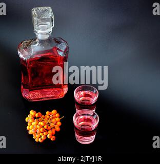 Hausgemachter Ebereschenlikör in zwei Gläsern und einer Flasche auf schwarzem Hintergrund, neben einem Haufen reifer Beeren. Nahaufnahme. Stockfoto