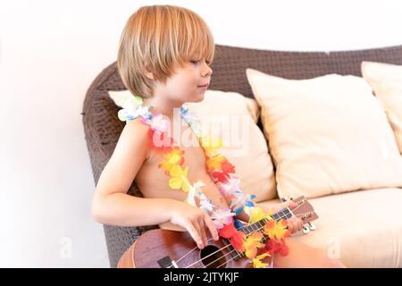 Kleiner glücklicher Junge spielt Gitarre oder Ukulele und sitzt draußen auf der Terrasse Stockfoto