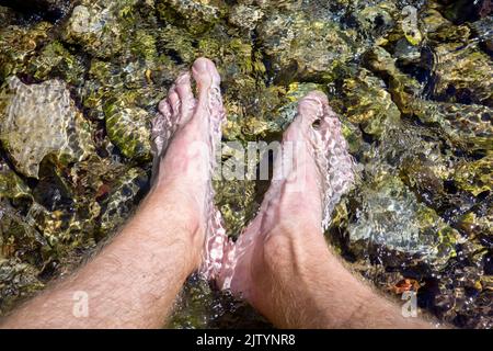 Männliche Füße in das klare Wasser eines Bergstroms eingetaucht. Abkühlung an einem heißen Tag. Stockfoto