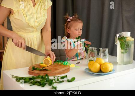 Frau hält Messer und schneiden Zitronen. Mama mit kleinen Tochter machen hausgemachte Limonade mit frischen gelben Zitronen und grüne Minze. Geringe Tiefe des Fiels Stockfoto