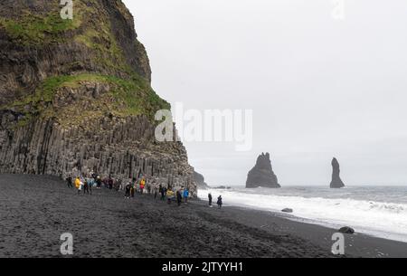 Touristen am Reynisfjara Strand, Klettern auf den Basaltsäulen und Ausweichen der Wellen, Island. Stockfoto