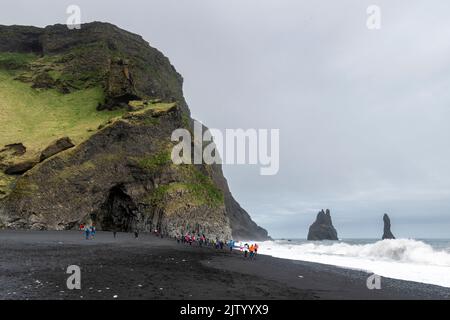 Touristen am Reynisfjara Strand, Klettern auf den Basaltsäulen und Ausweichen der Wellen, Island. Stockfoto