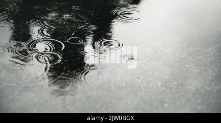 Ein schwarz-weißes Bild einer tiefen Pfütze in der Stadt, in die Regentropfen fallen und Kreise auf dem Wasser hinterlassen. Schlechtes, trauriges Wetter. Stockfoto