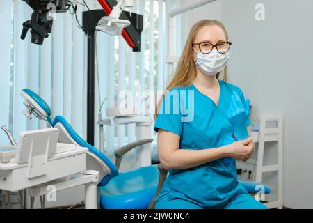 Glückliche kaukasische weibliche Zahnärztin in blauem Labormantel und schützender Gesichtsmaske, die mit einem hübschen Lächeln posiert und in ihrer Zahnarztpraxis auf die Kamera schaut Stockfoto