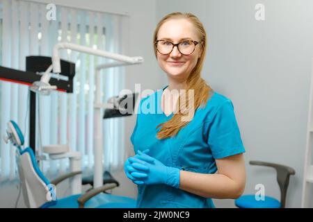 Glückliche kaukasische weibliche Zahnärztin in blauem Labormantel und schützender Gesichtsmaske, die mit einem hübschen Lächeln posiert und in ihrer Zahnarztpraxis auf die Kamera schaut Stockfoto