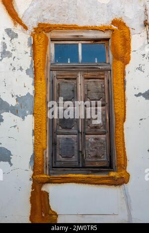 Alte rustikale Holzlamelle, die ein Fenster in einer weiß getünchten Wand eines traditionellen griechischen Gebäudes bedeckt, schäbiges, schickes, verwittertes Fenster in einer weiß getünchten Wand. Stockfoto