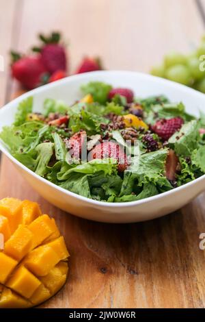 Salat aus Salat, Mango, Erdbeere, Nüssen und Samen. Serviert in einer weißen Schüssel auf einem Holztisch. Gesunde Mahlzeit in einem Restaurant Stockfoto