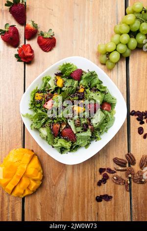 Salat aus Salat, Mango, Erdbeere, Nüssen und Samen. Serviert in einer weißen Schüssel auf einem Holztisch. Gesunde Mahlzeit in einem Restaurant Stockfoto