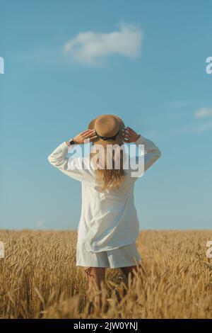 Rückansicht einer jungen Frau in einem weißen Kleid und einem Strohhut auf einem Weizenfeld im Sommer. Stockfoto